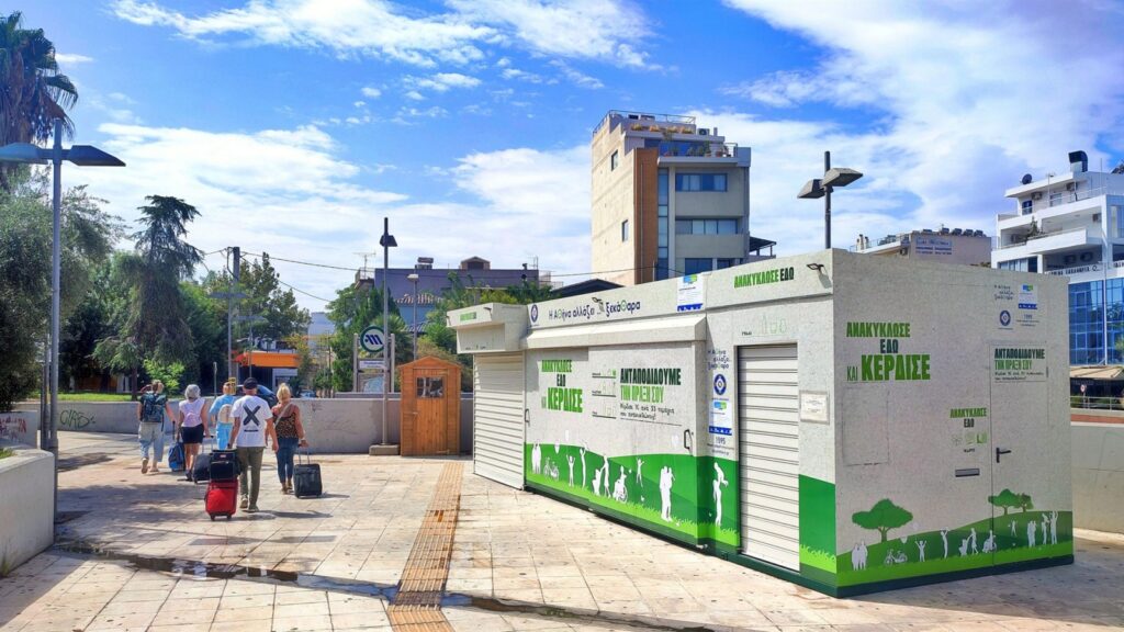Σταθμός «Σπιτιού Ανακύκλωσης» σε αστικό χώρο με φόντο σύμβολα ανακύκλωσης και γραφικά που υποδηλώνουν οικονομικές απώλειες και ευρωπαϊκό έλεγχο